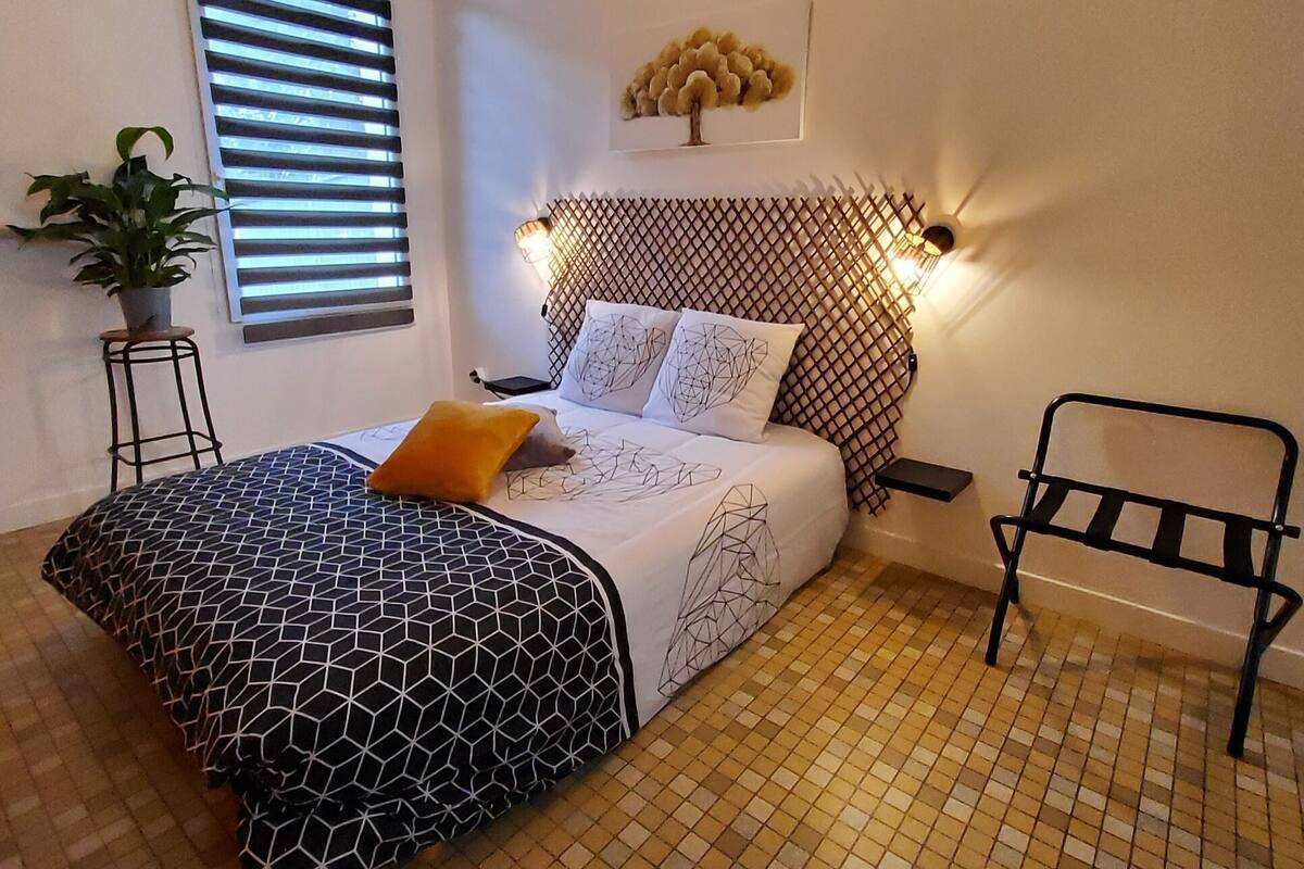 A bedroom with a geometric-patterned bedspread, a potted plant, a wall-mounted light, and a tiled floor.