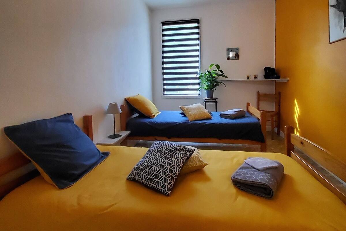 A room with two beds, a window with blinds, a plant, and a shelf with items.