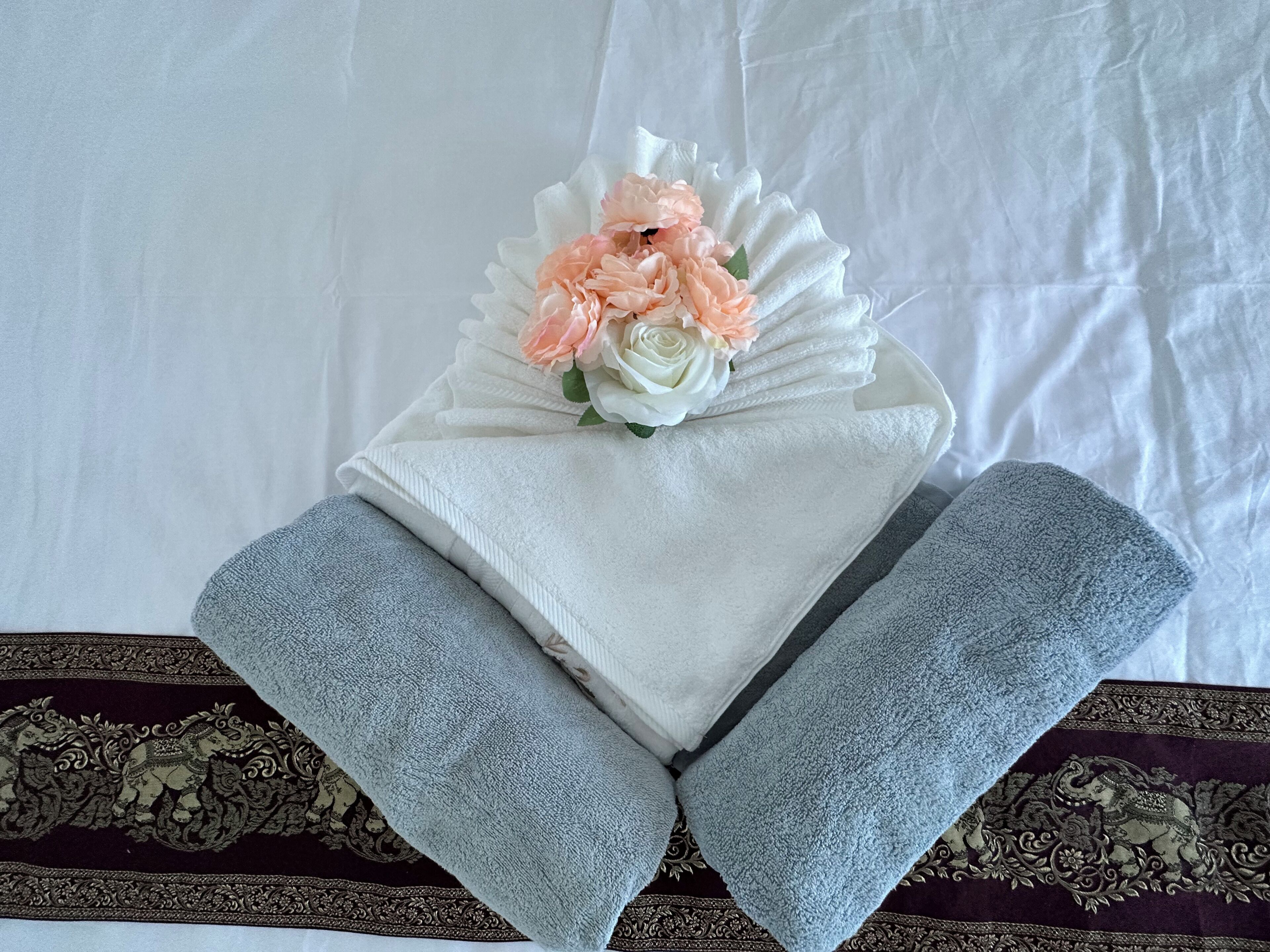 Una toalla blanca doblada con un arreglo floral encima, sobre una cama con sábanas grises y una colcha estampada.