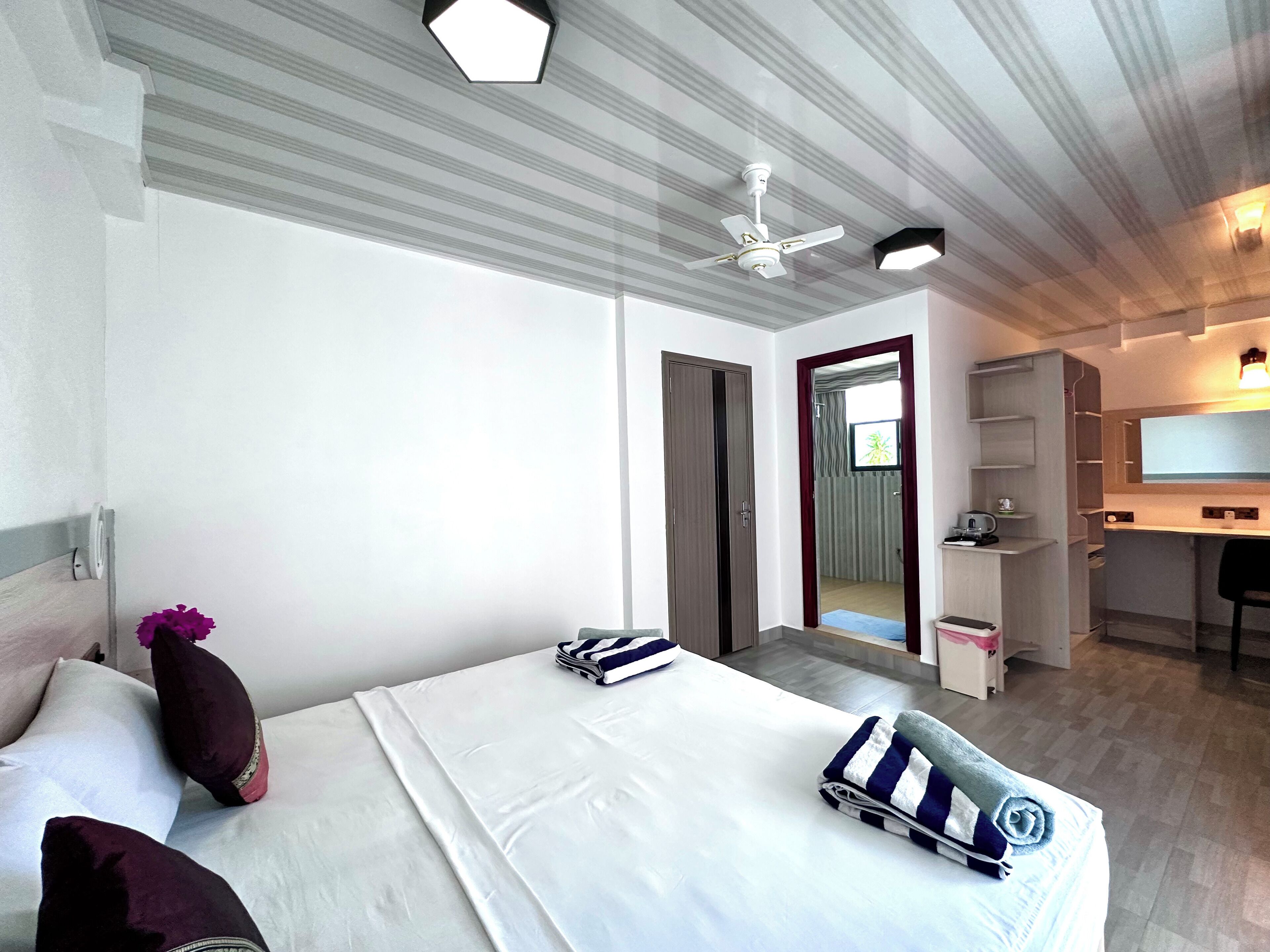 Quarto de hotel com cama, escrivaninha, cadeira, guarda-roupa e ventoinha no teto.