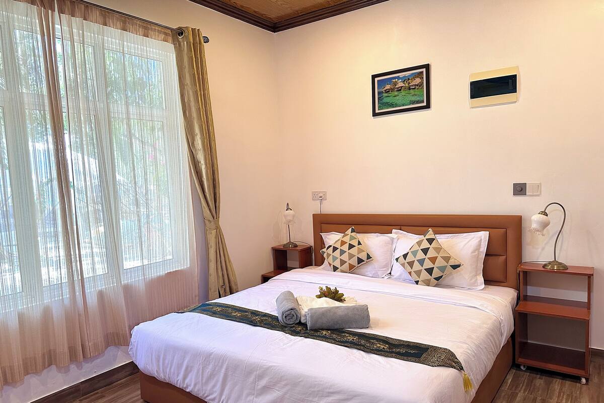 Cama bem arrumada com lençóis brancos e almofadas decorativas, mesa de cabeceira com abajur e janela com cortinas finas.