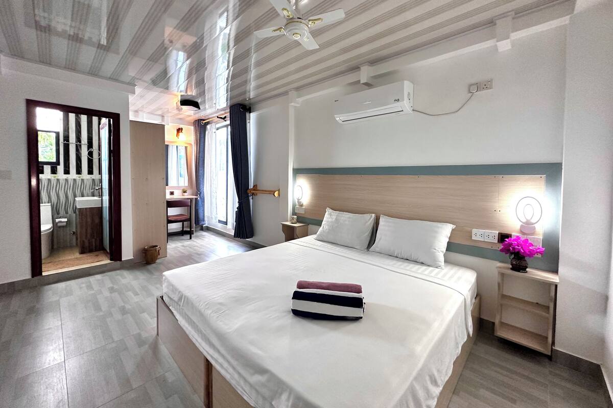 Quarto de hotel com uma cama grande, uma mesa de cabeceira com luminária, ventilador de teto e ar condicionado.