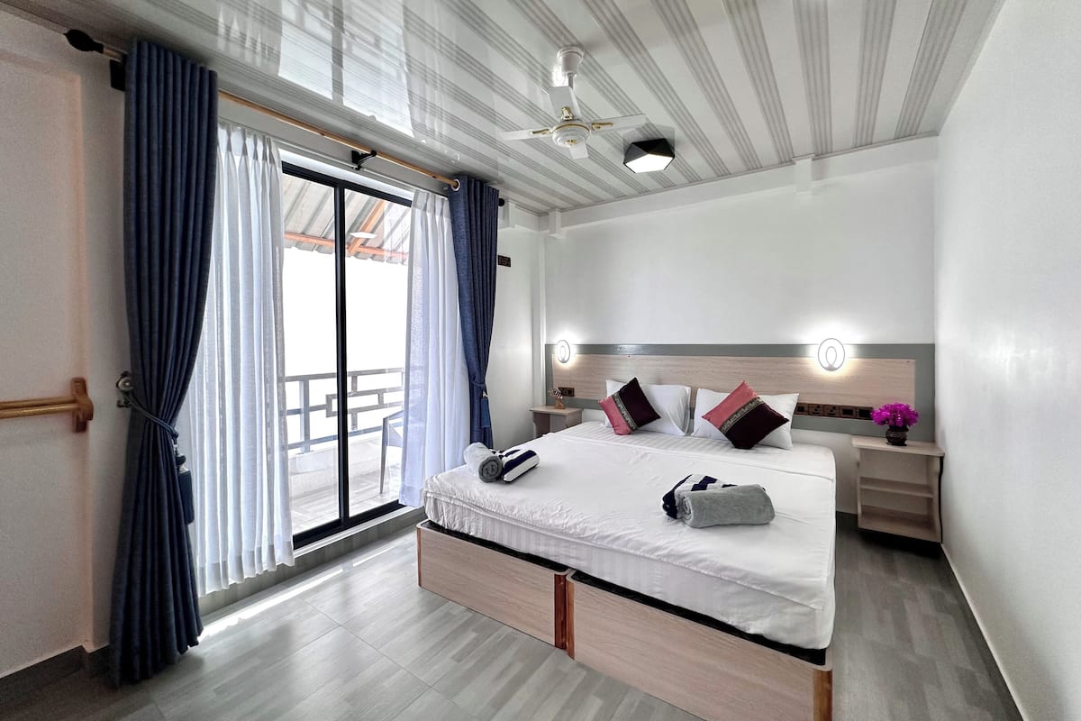 Quarto de hotel moderno com uma cama grande, ventilador de teto e vista para a varanda externa.