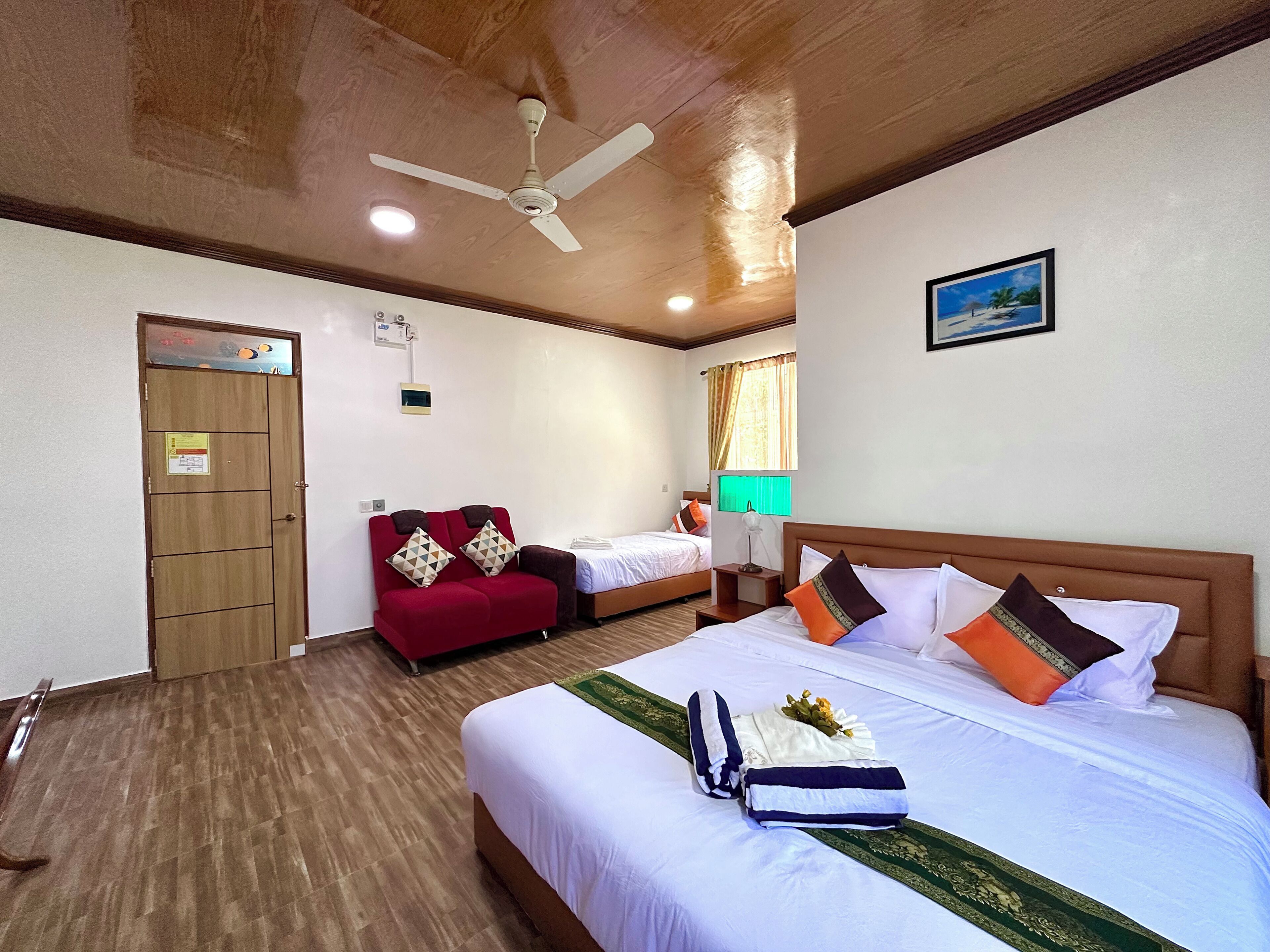 Habitación de hotel con techo de madera, cama con ropa blanca y varios almohadones, sofá rojo con cojines estampados, mesita de noche con lámpara y ventana con cortinas.