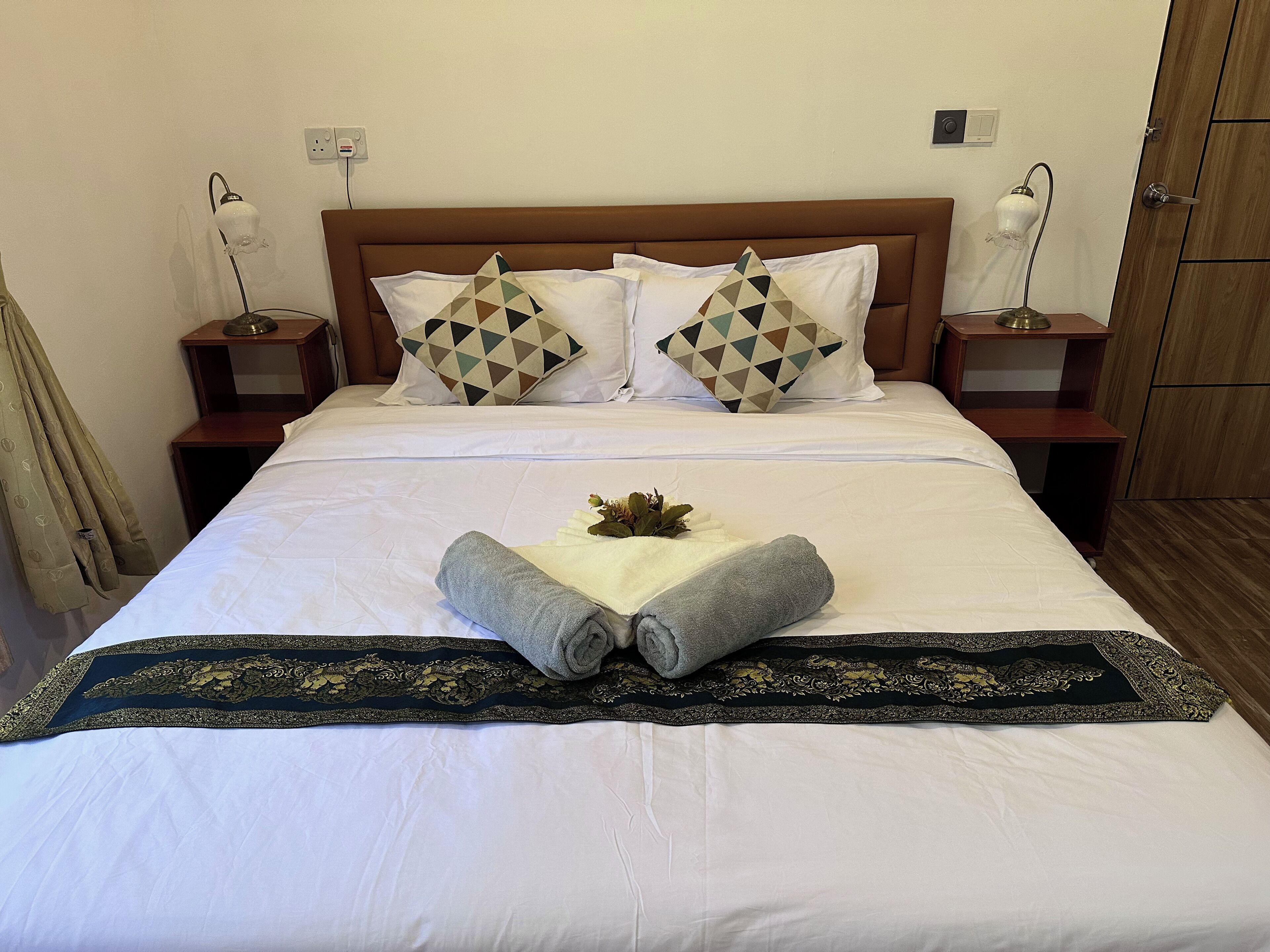 Una cama bien tendida con ropa blanca, dos almohadas decorativas, una toalla doblada y un cabecero decorativo.