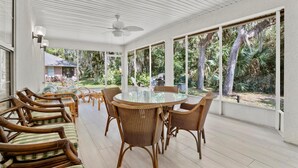 Een overdekt terras met een ronde glazen tafel en rieten stoelen, grote ramen met uitzicht op bomen en een huis.