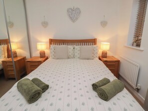 A neatly made bed with a patterned bedspread, two bedside lamps, heart-shaped wall decorations, and a window with blinds. - Brucap Cottage (Chathill)