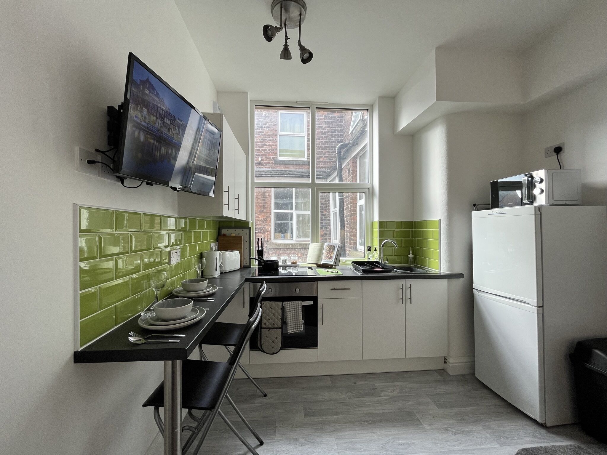 A compact kitchen with a TV mounted above the counter, a window with a view of a brick building, and a white refrigerator.