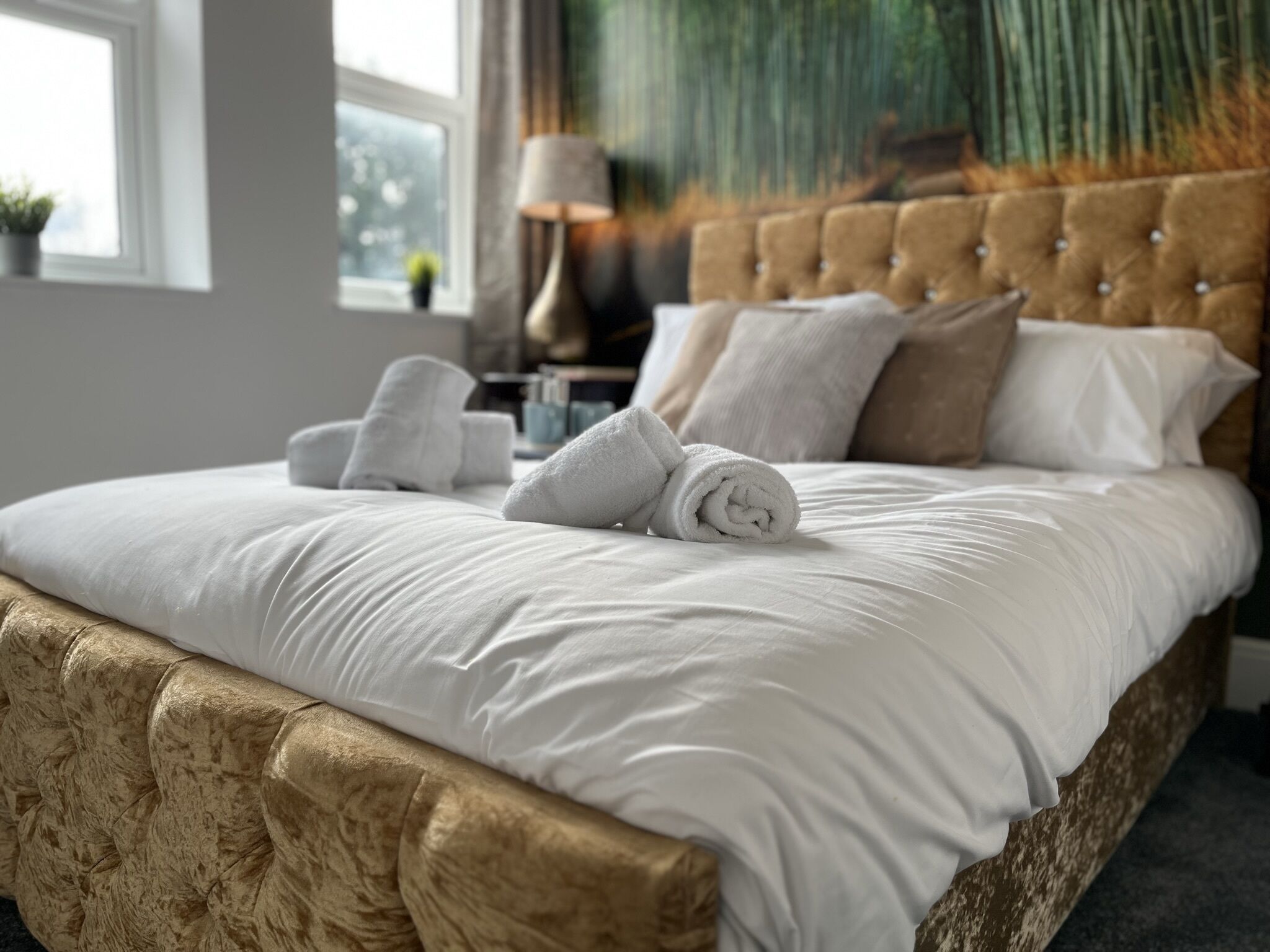 A neatly made bed with white linens and rolled towels, a tufted headboard, and a window with a view of greenery.