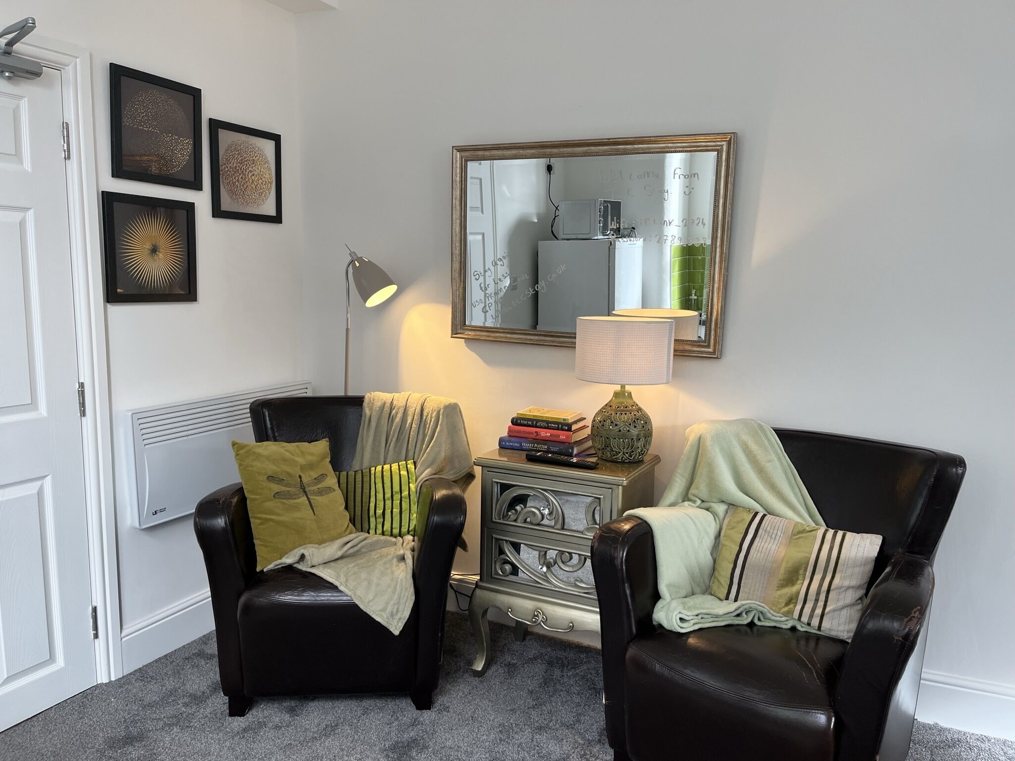 A room with two black leather chairs, a small side table with books and a lamp, a mirror on the wall, and framed artwork.