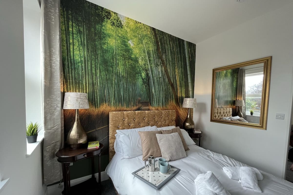 Habitación de hotel con un amplio mural con tema de bosque, una cama bien hecha con ropa blanca, una mesita de noche con lámpara y un espejo en la pared.
