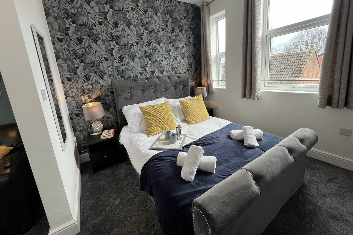 Habitación de hotel con una cama grande, un banco tapizado en gris, una mesita de noche con lámpara y papel tapiz floral.