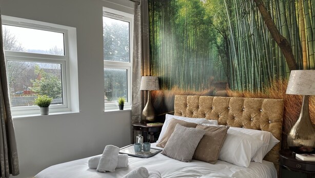 Una cama bien hecha con sábanas blancas y toallas enrolladas, un ventanal con vistas a árboles y un mural en la pared con un bosque de bambú.