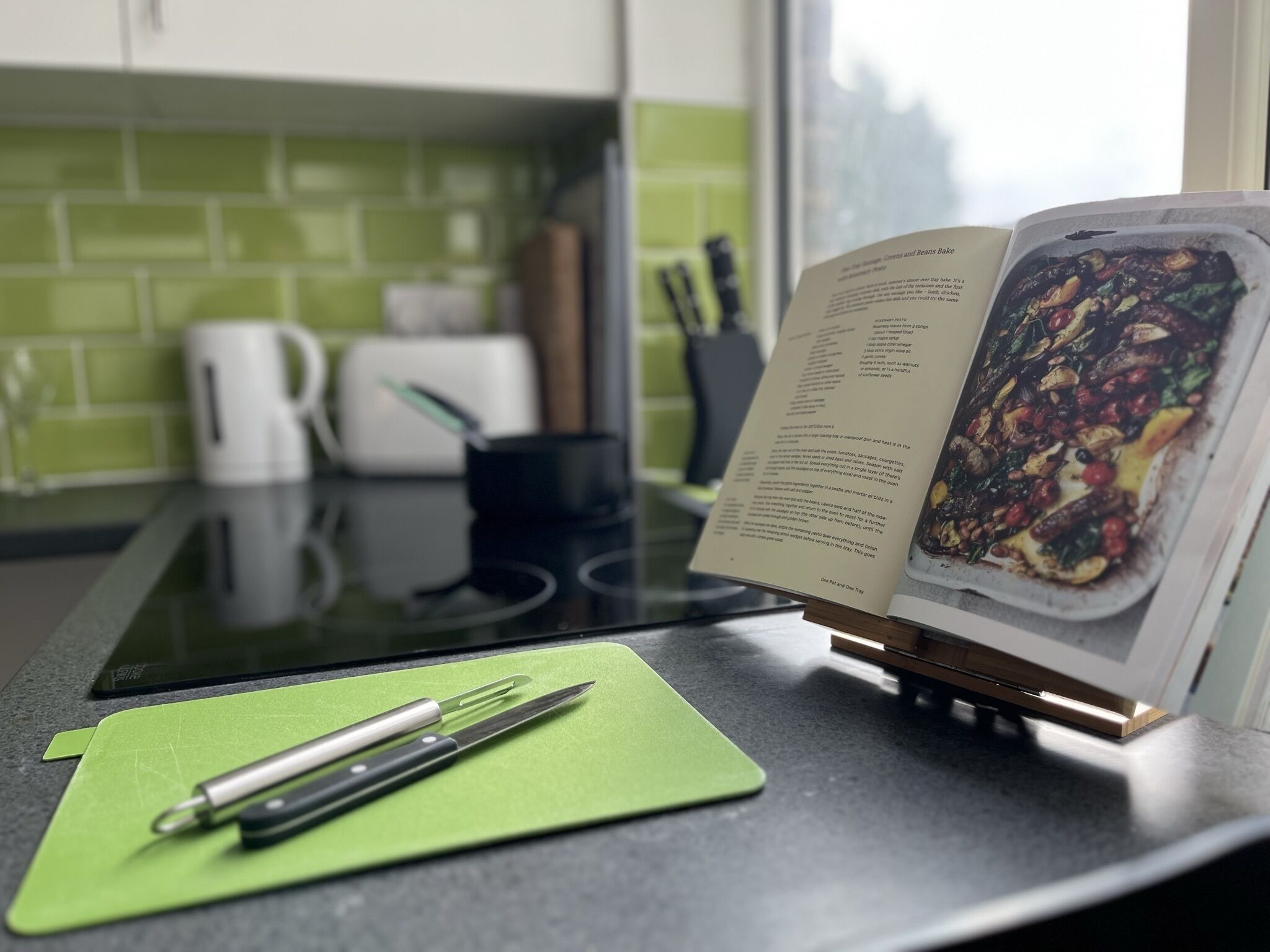 Un piano di lavoro in cucina con un tappetino verde da taglio, una penna e un libro di ricette aperto che mostra un piatto con verdure arrostite.