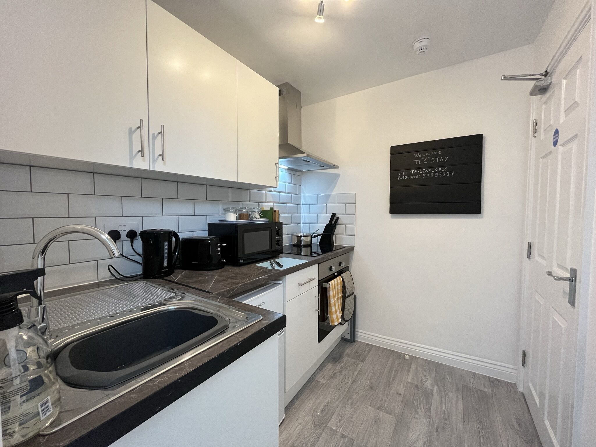 A compact kitchen with white cabinets, a black microwave, a kettle, and a sink. There's a chalkboard with Wi-Fi and password information on the wall.