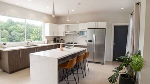 Een moderne keuken met een groot eiland, barkrukken, een roestvrijstalen koelkast en uitzicht op groen door grote ramen.