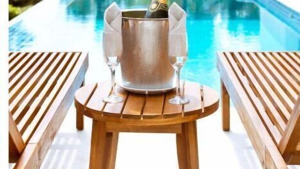 Een houten tafel met een champagnekoeler en glazen, met uitzicht op een zwembad en een tropische achtergrond.