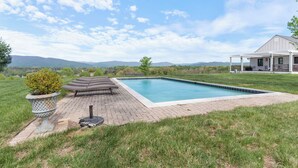 A rectangular swimming pool with a brick deck, a covered patio area, and a house in the background. - Rustic Bailey's Retreat Cabin Near Wineries! (Charlottesville)