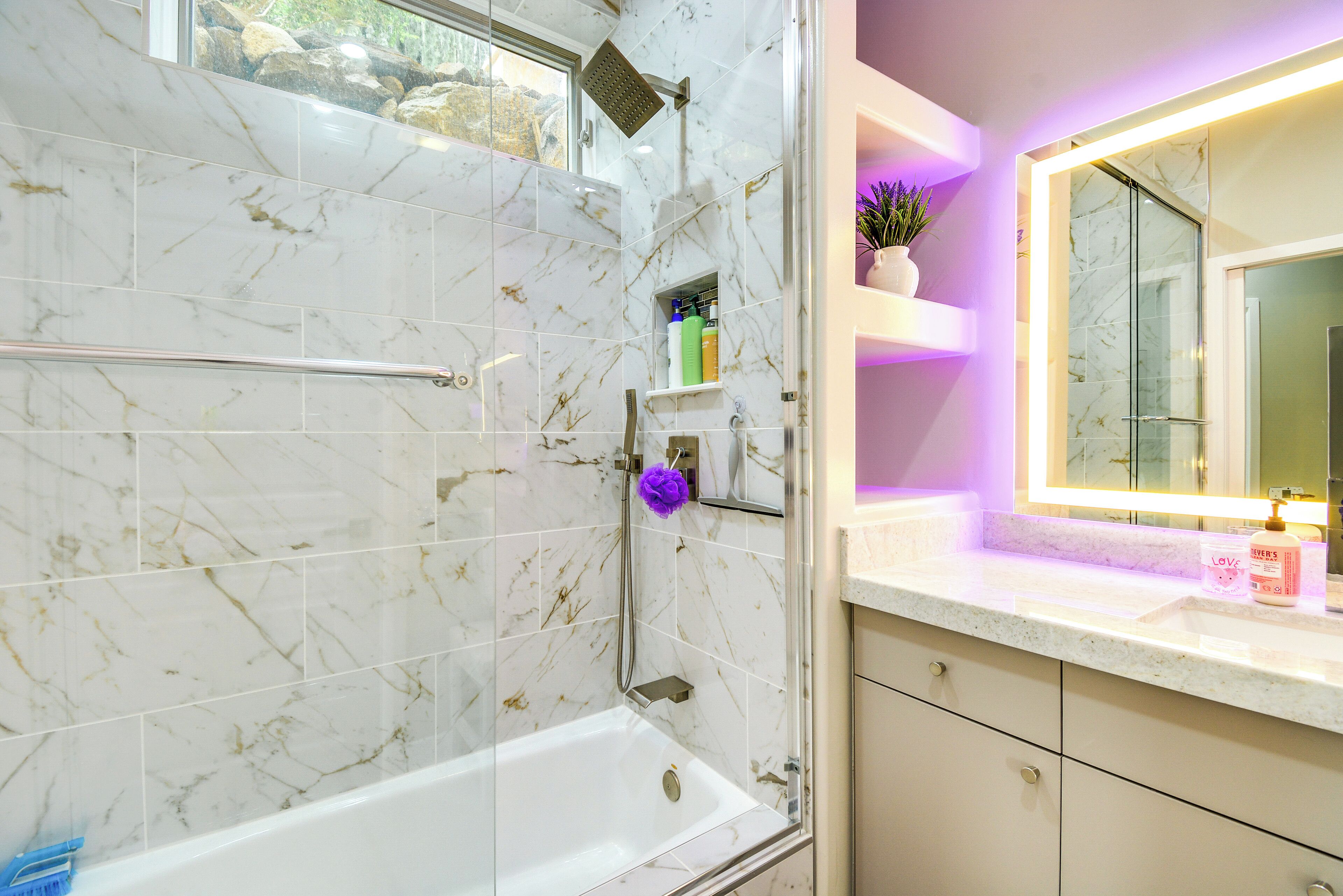 A modern bathroom with a glass-enclosed shower, marble tiles, a vanity with a mirror, and a small shelf with toiletries.