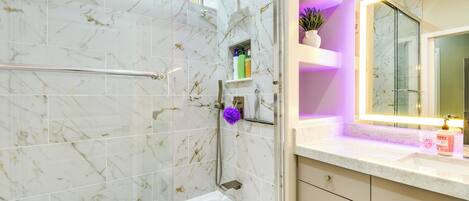 A modern bathroom with a glass-enclosed shower, marble tiles, a vanity with a mirror, and a small shelf with toiletries.