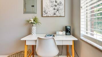 Un bureau blanc avec une chaise blanche, un vase de fleurs, un tableau abstrait encadré et une fenêtre avec des stores.