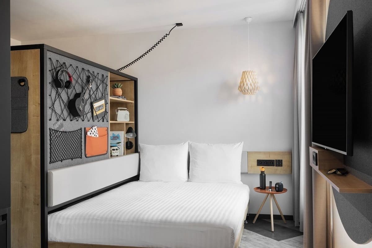 A modern hotel room with a bed, a TV, a desk with a lamp, and a wall-mounted shelf with personal items.