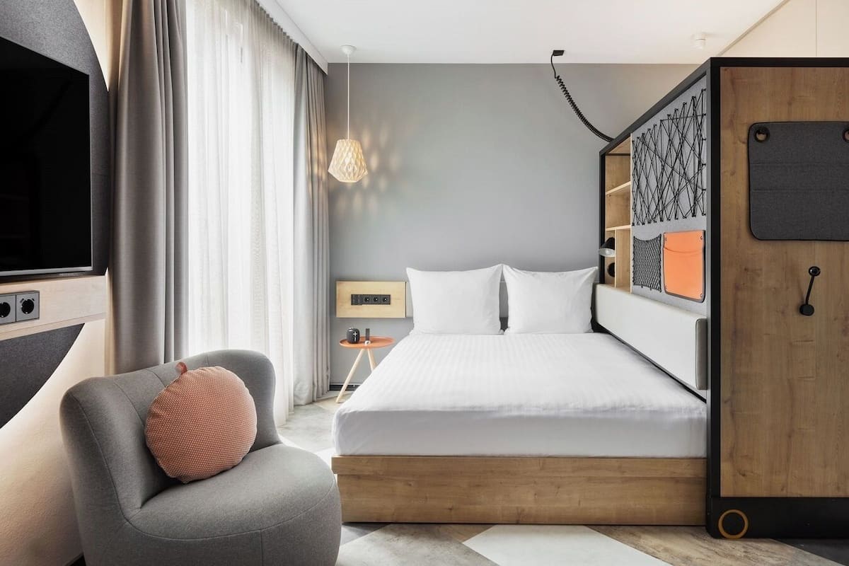 A modern hotel room with a bed, a gray armchair, a TV, and a wardrobe.