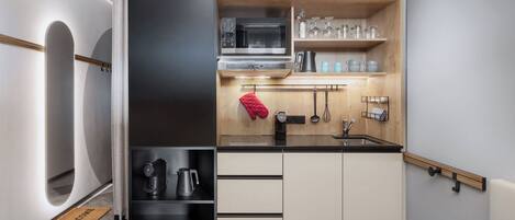 A compact kitchen with a microwave, kettle, and coffee maker, a wooden shelf with glassware, and a black countertop.