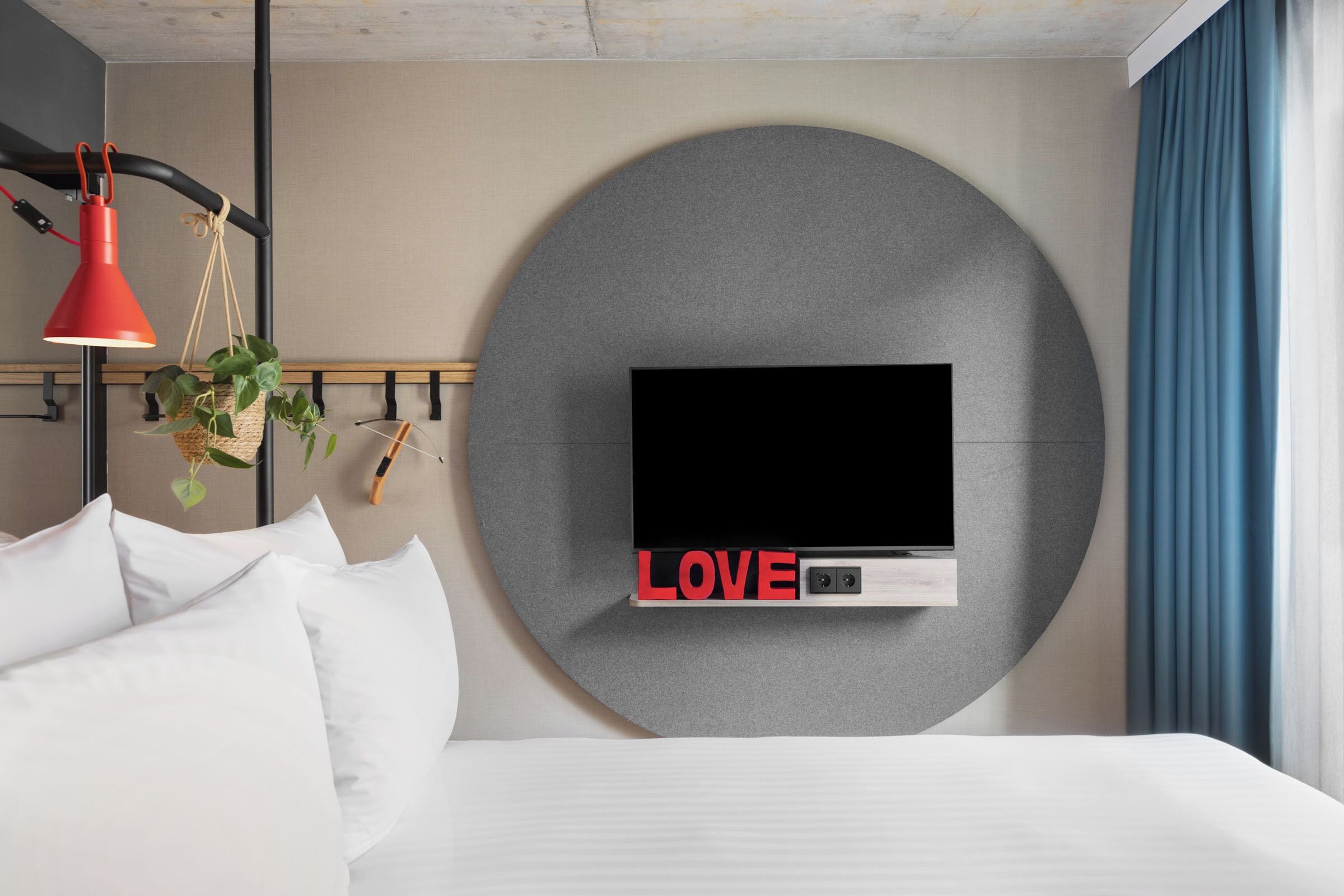 Ett modernt hotellrum med en stor säng, en väggmonterad TV, en röd hänglampa och en rund väggdekoration med ordet ”KÄRLEK”.