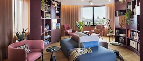 A modern living room with a sofa, chairs, a bookshelf, and a large window with a view of a building.