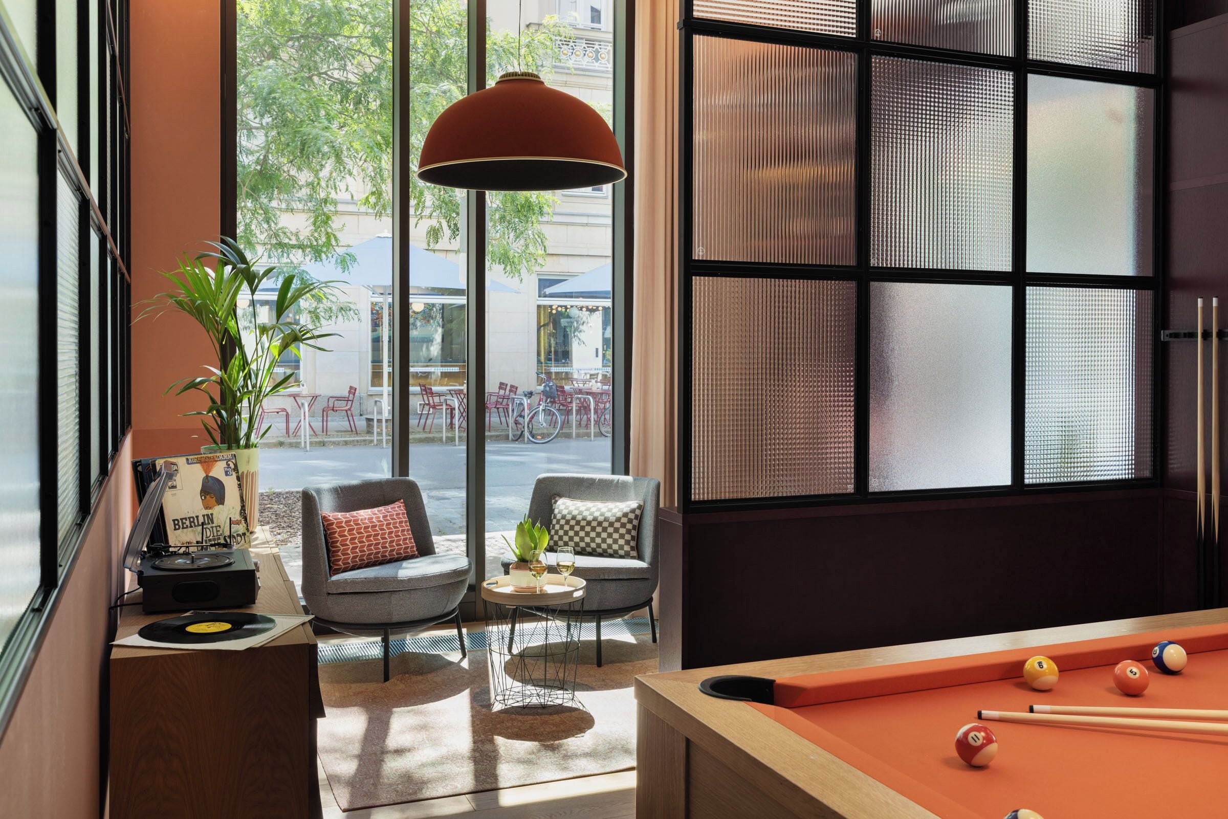 Una habitación con mesa de billar, sofá, una mesita con una planta y ventana con vistas a bicicletas afuera.