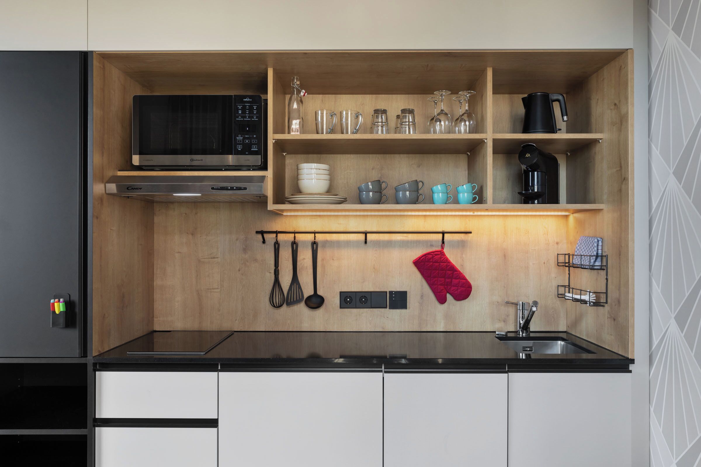 A compact kitchen with a microwave, oven, and sink. There are shelves with dishes and glasses, and a red oven mitt hanging.