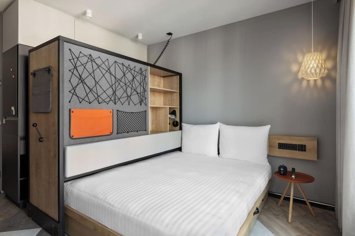 A modern bedroom with a bed, a small bedside table, a wall-mounted shelf, and a hanging light fixture.
