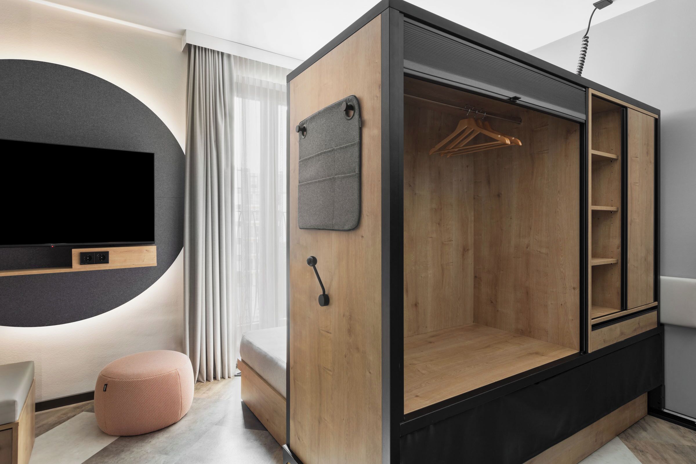 A modern room with a built-in TV, a wooden wardrobe with a hanging space, and a small ottoman.