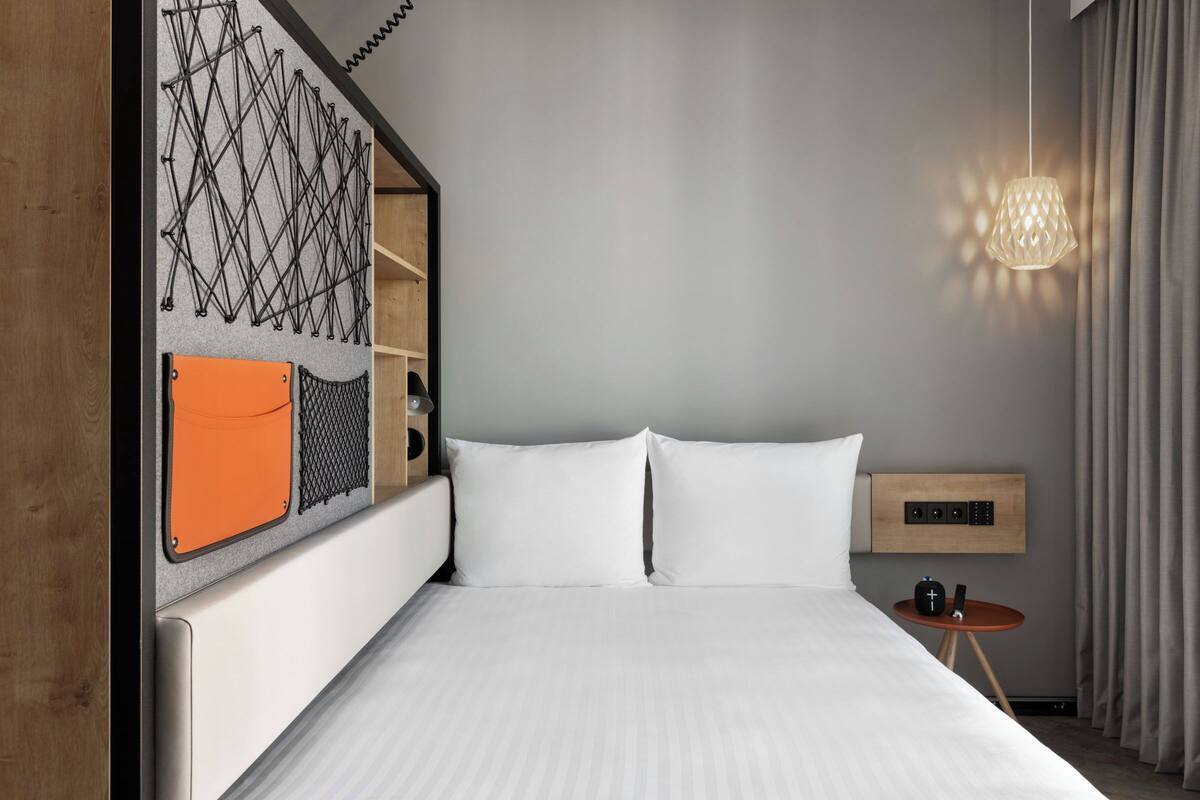 A modern hotel room with a neatly made bed, a wall-mounted shelf with a decorative item, a small table with a lamp, and a window with curtains.