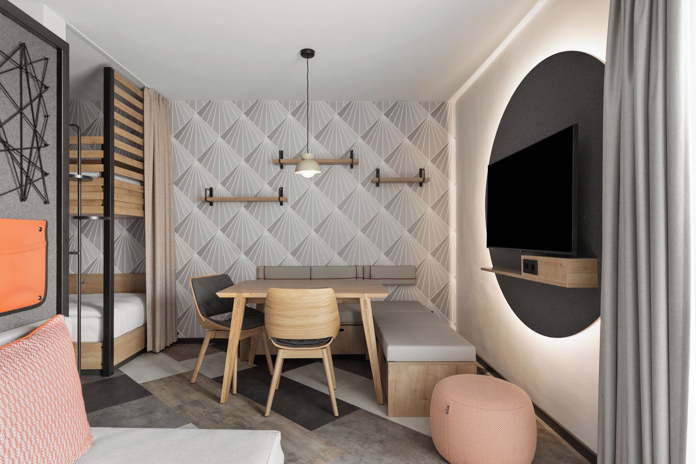 A compact room with a bed, a desk, a chair, a round ottoman, and a TV mounted on the wall.