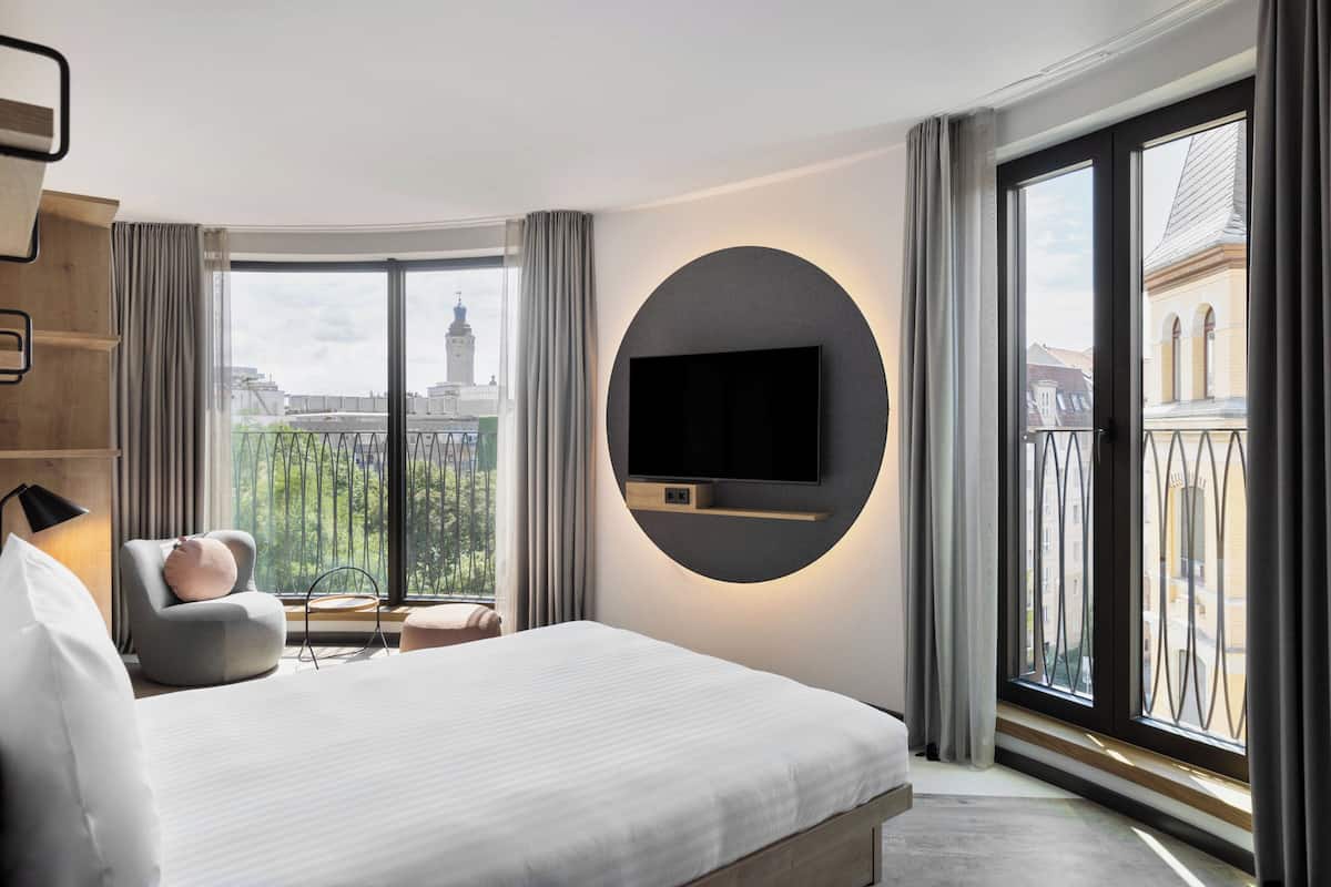 A modern hotel room with a large bed, a mounted TV, a view of the city through large windows, and a balcony with a railing.