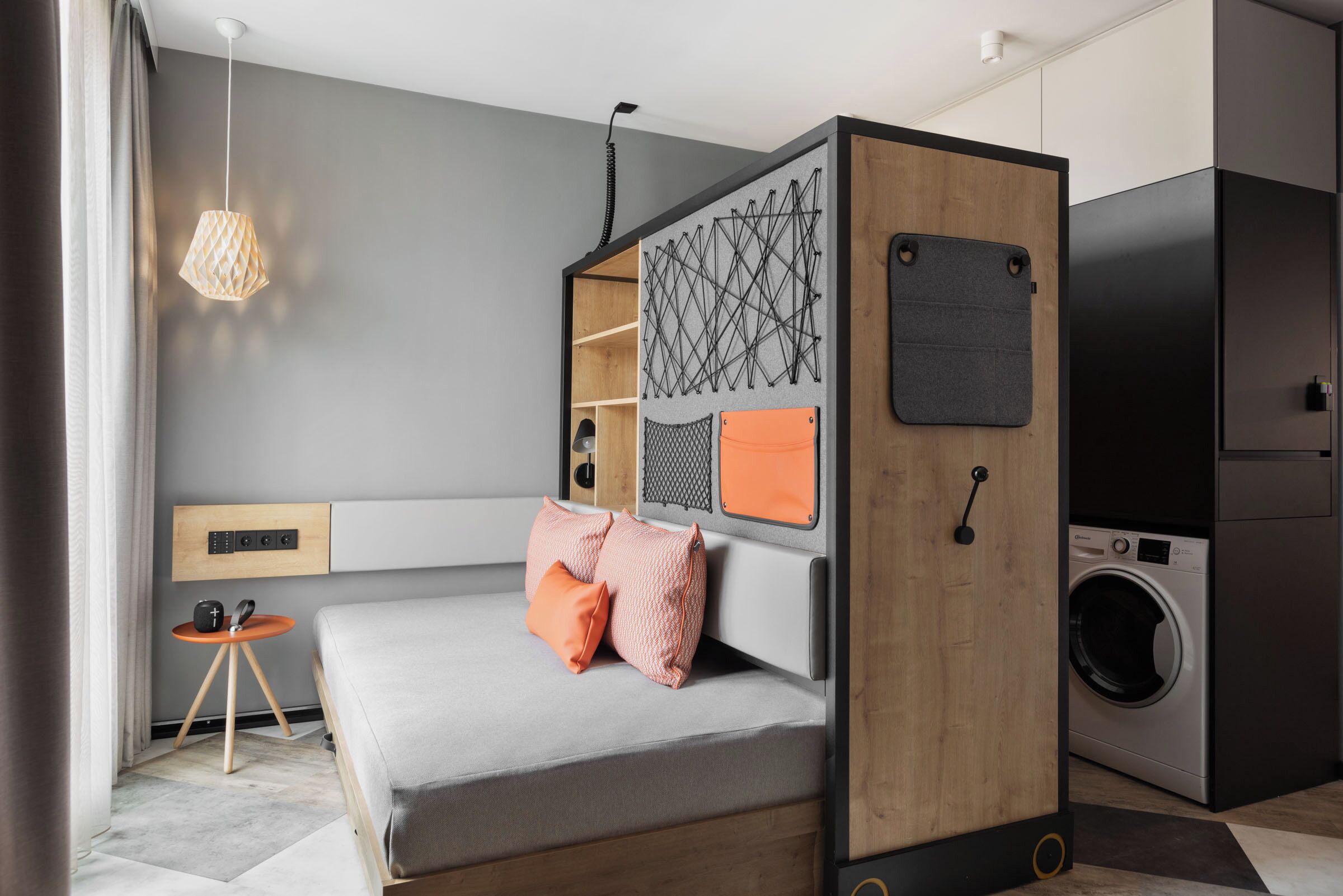 A compact bedroom with a bed, a small table, a washing machine, and a hanging light fixture.