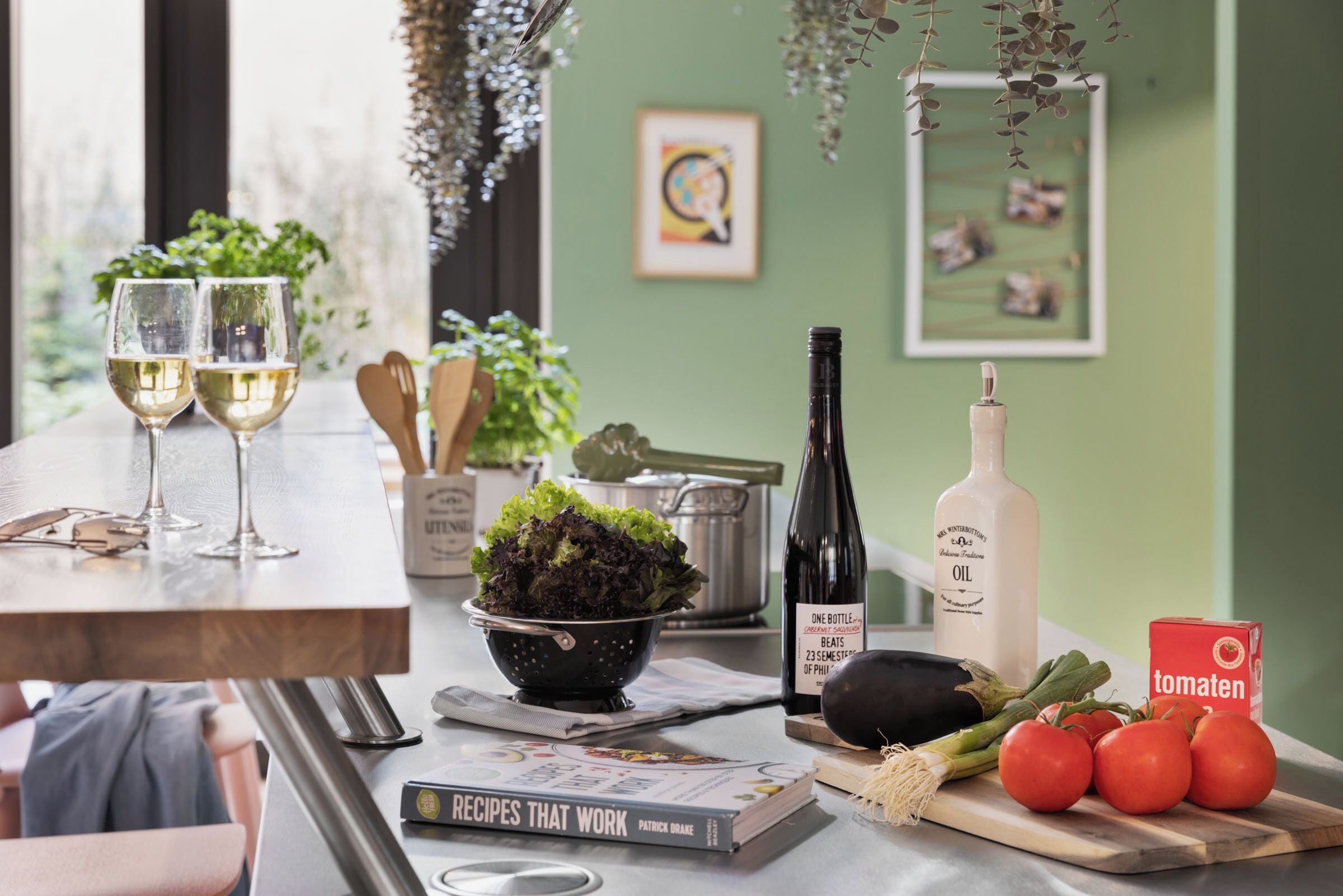 A kitchen counter with two wine glasses, a bowl of salad, a bottle of oil, a book titled 'Recipes That Work,' and fresh tomatoes.