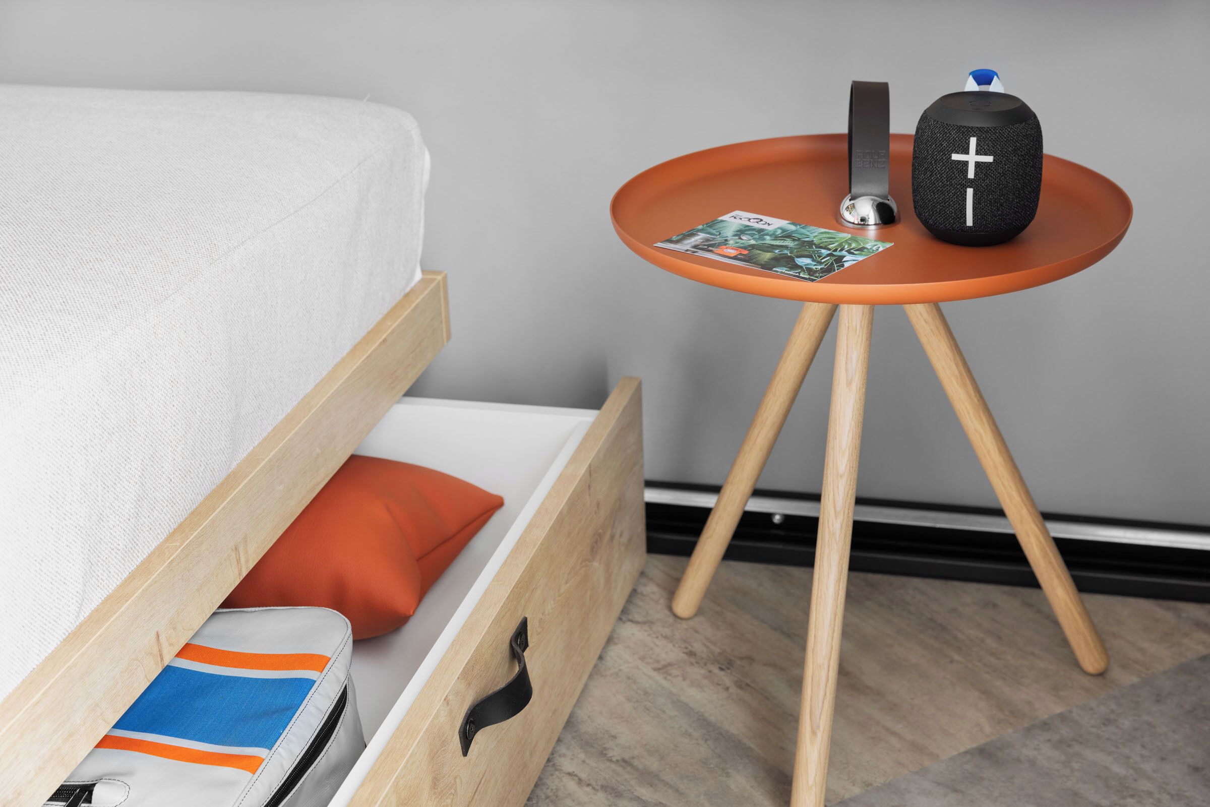 Una cama con colchón blanco y estructura de madera, un pequeño cojín naranja, un cajón con una bolsa a rayas, y una mesa con una revista, un altavoz negro y un objeto azul.