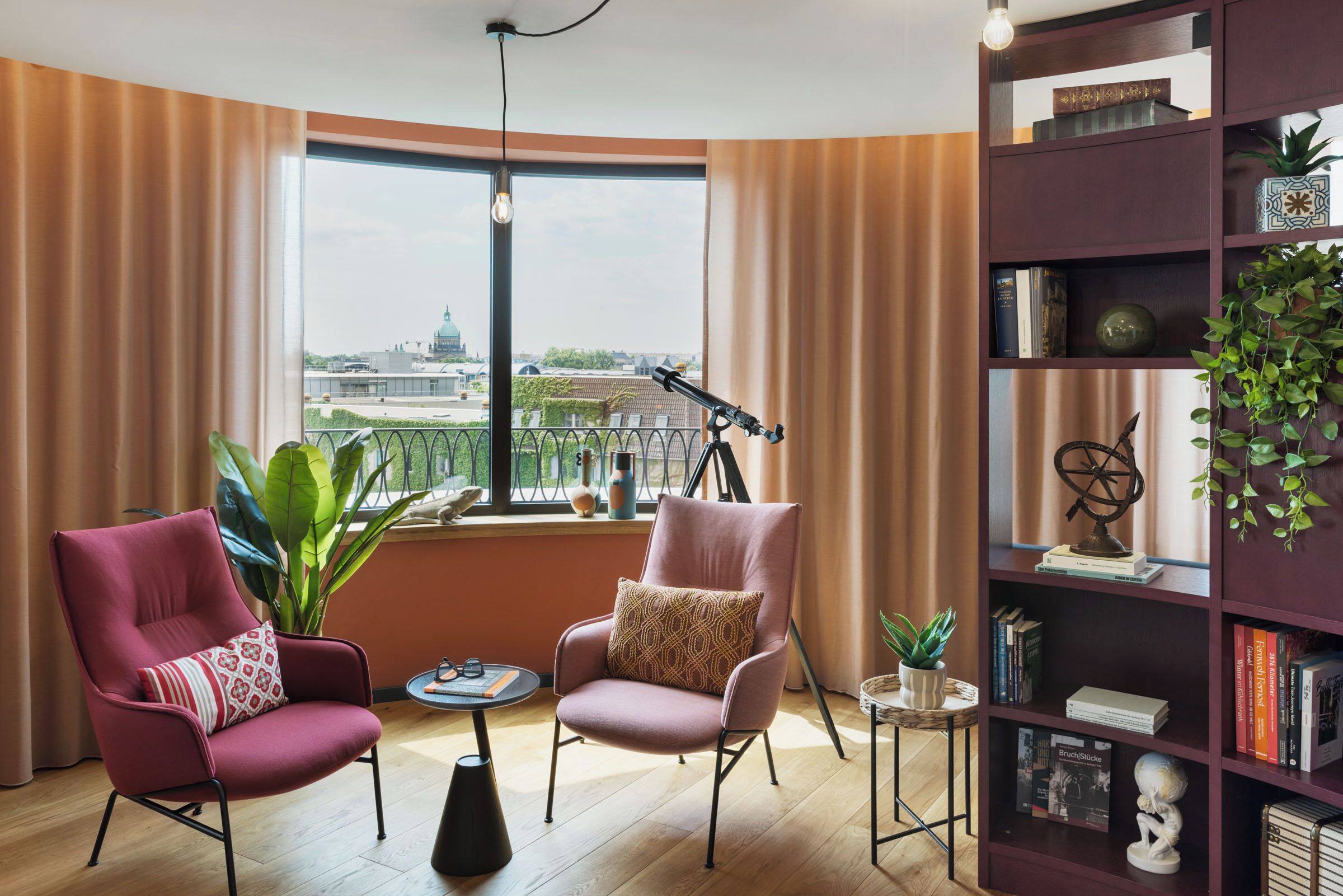 A room with two armchairs, a small table, a bookshelf, and a large window with a view of a cityscape.