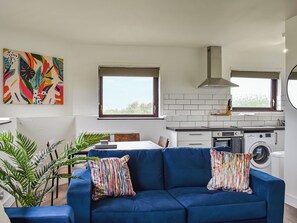 Uma sala de estar moderna com um sofá azul, duas almofadas coloridas, um grande quadro abstrato e vista para a cozinha, onde se vê uma máquina de lavar.