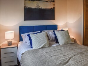 Un lit bien fait, avec une tête de lit bleue, des oreillers blancs et bleus, et une couverture grise. On y trouve une table de chevet avec une lampe et un petit objet décoratif. Un tableau est accroché au-dessus du lit.