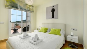 A neatly made bed with white linens and green pillows, a bedside table with a lamp, and a window with a view of greenery. - Terrazas de Alcaidesa 2437 Penthouse (San Roque)