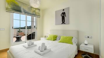 Una cama bien hecha con sábanas blancas y almohadas verdes, una mesita de noche con una lámpara, y una ventana con vistas a zonas verdes.