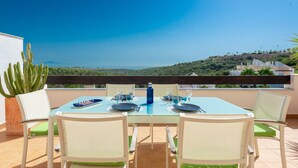 A dining table with chairs on a terrace with a view of a hill and ocean. - Terrazas de Alcaidesa 2437 Penthouse (San Roque)