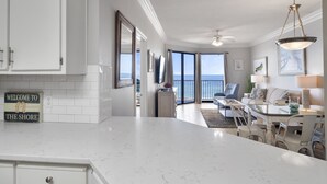 A spacious living area with a large window overlooking the sea, a dining area with white furniture, and a kitchen with white cabinets.