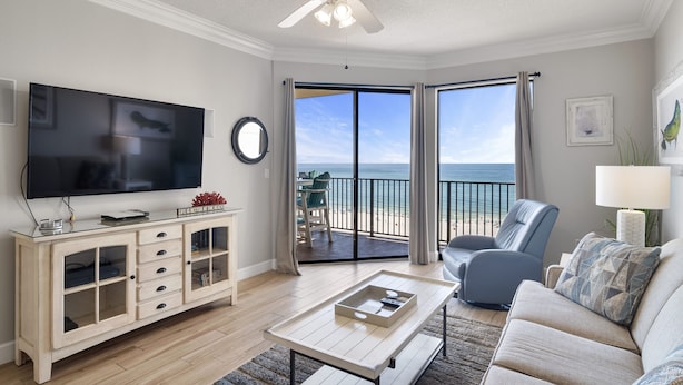 A living room with a television, a ceiling fan, a coffee table, a sofa, and a view of the beach through large sliding doors.