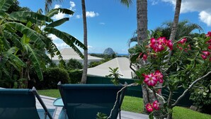 A deck with two green chairs, a potted plant with pink flowers, and a view of a building with a white roof.