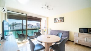 Un salon moderne avec une table ronde en bois, des chaises noires, un canapé, un téléviseur et un balcon offrant une vue sur la ville.